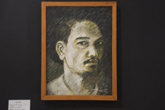 Framed charcoal portrait of a man with short hair and mustache, on a dark gallery wall.