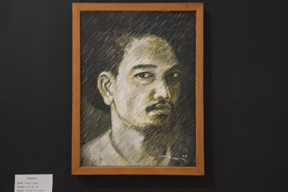 Framed charcoal portrait of a man with short hair and mustache, on a dark gallery wall.