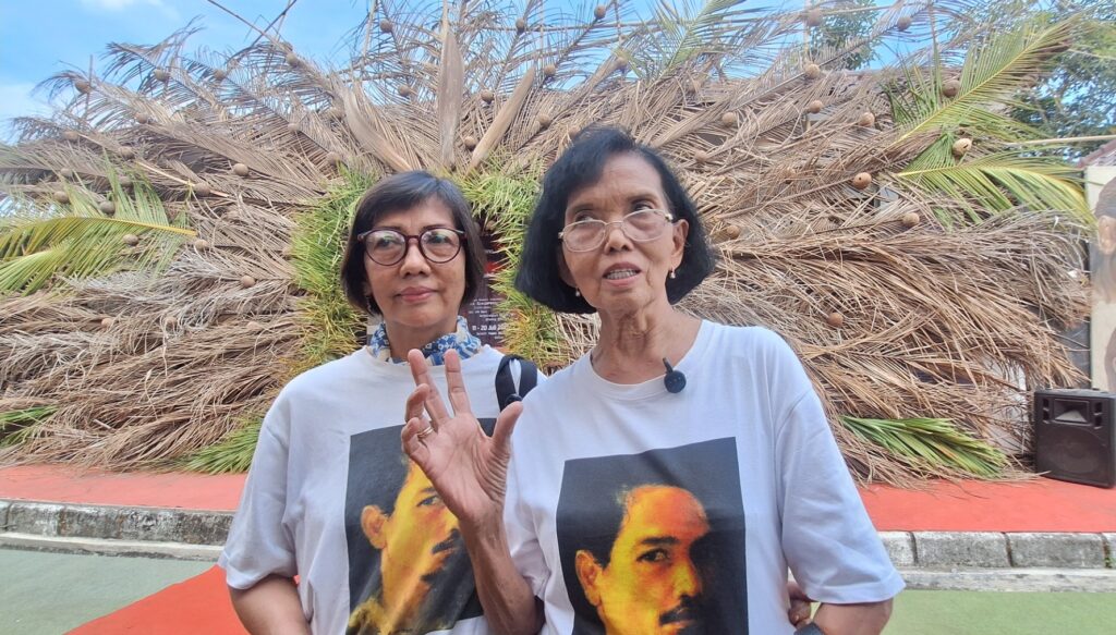 Screenshot_20260716_172125_Gallery - Mabur.co Two women posing outdoors in front of a giant dried palm sculpture, wearing matching white shirts with a large portrait image; one shows a peace sign with fingers partially raised.