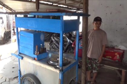 Street vendor beside a blue wooden cart with bottles on the counter, man in a cap and casual clothes nearby.