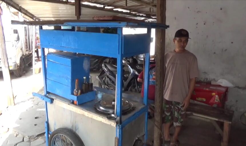 Street vendor beside a blue wooden cart with bottles on the counter, man in a cap and casual clothes nearby.
