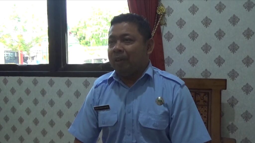 Middle-aged man in a light blue uniform shirt with a name badge sits indoors by a window.