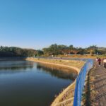People walking on a blue railing-lined promenade beside a calm river under a clear blue sky.