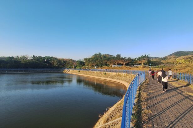 People walking on a blue railing-lined promenade beside a calm river under a clear blue sky.