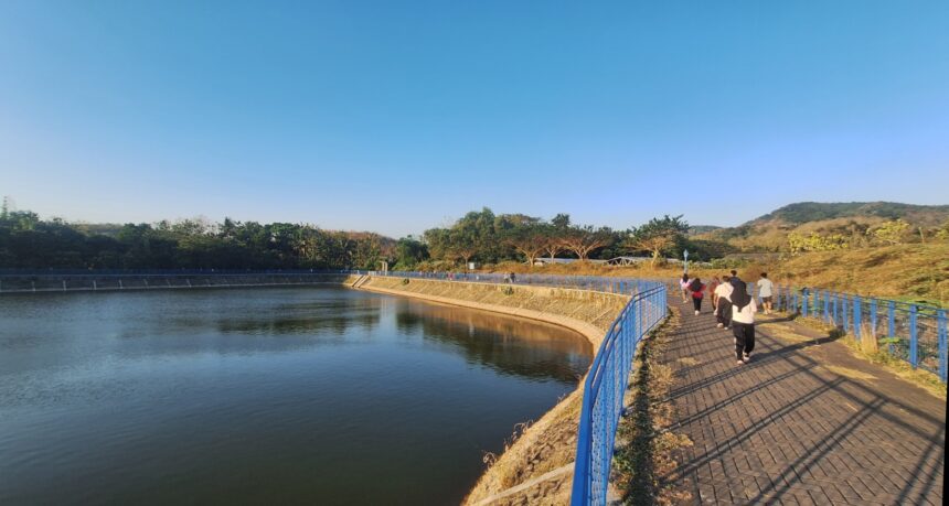 People walking on a blue railing-lined promenade beside a calm river under a clear blue sky.