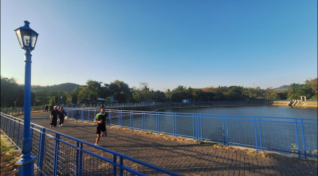 Blue lamppost overlooking a brick walkway along a blue railing beside a calm river or lake, with joggers in the distance