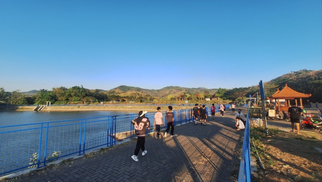 People walk along a paved riverside path with blue railings, water on the left and hills in the distance, a pavilion on the right.