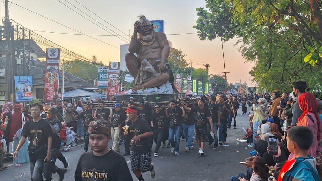 Screenshot_20260717_183110_Gallery - Mabur.co Street parade with a huge seated demon statue on a float, crowds lining the road and banners in the background.