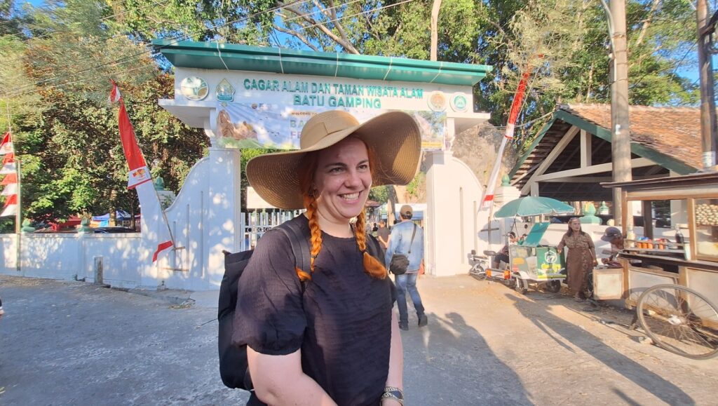 Screenshot_20260717_183356_Gallery - Mabur.co Smiling woman with long orange braids and a wide straw hat stands in front of Batu Gamping Nature Reserve entrance. Behind her are trees and a market area.