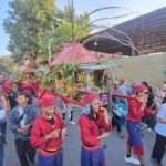 Parade scene with performers in red outfits carrying a plant-filled float through a crowded street as spectators film and watch.