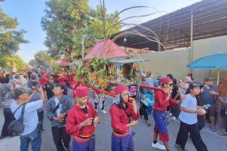 Parade scene with performers in red outfits carrying a plant-filled float through a crowded street as spectators film and watch.