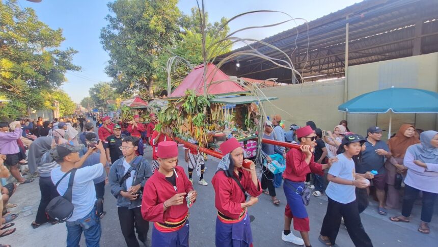 Parade scene with performers in red outfits carrying a plant-filled float through a crowded street as spectators film and watch.
