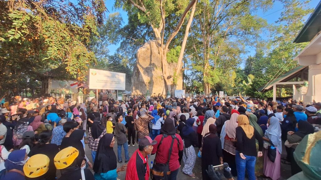 Screenshot_20260717_183635_Gallery - Mabur.co Large crowd of people gathered outdoors around a tall rock monument and banners, with trees and a building to the sides.