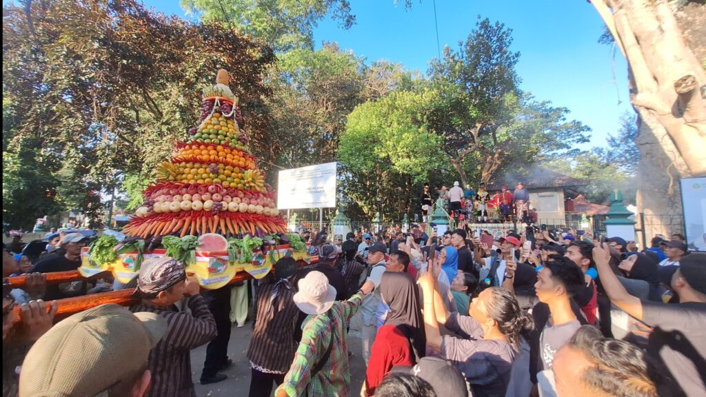 Colorful fruit pyramid float carried by a crowd during a street festival, with people taking photos under a clear blue sky.
