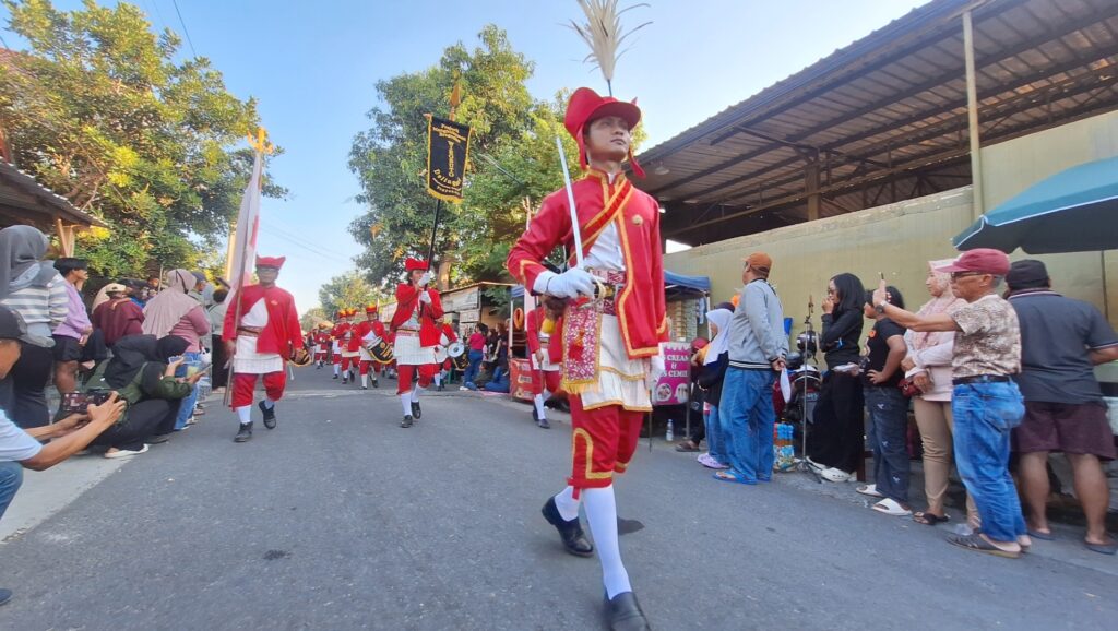 A man in a red ceremonial uniform leads a parade down a street with cheering spectators on both sides.