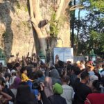 Large crowd gathered in front of a massive rock formation, with people raising hands toward a central notice board/signage and a green fence behind them.