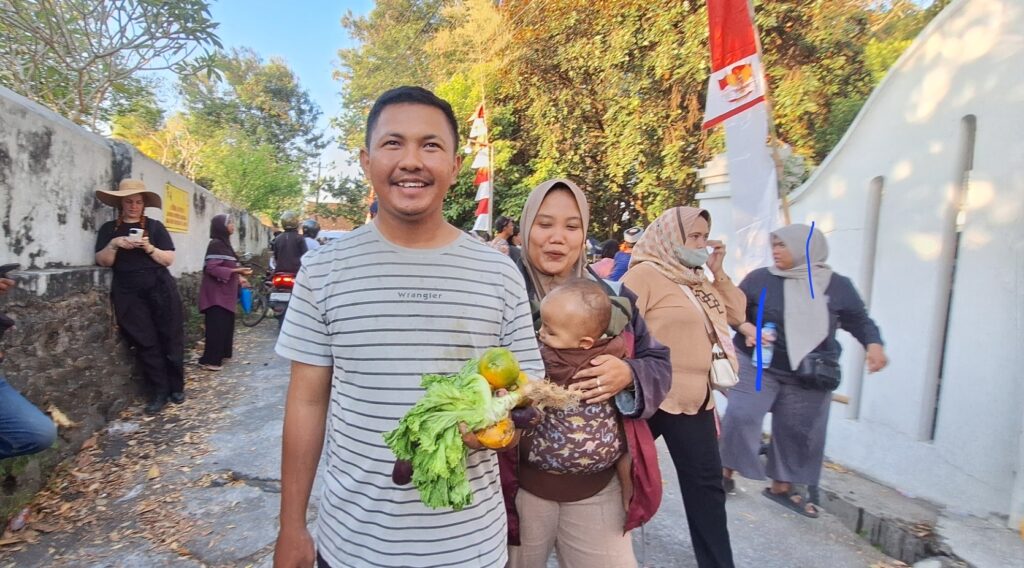 Screenshot_20260717_214200_Gallery - Mabur.co Man with striped shirt smiles at camera, holding fresh greens and a baby carrier with leafy vegetables in a sunny street scene.