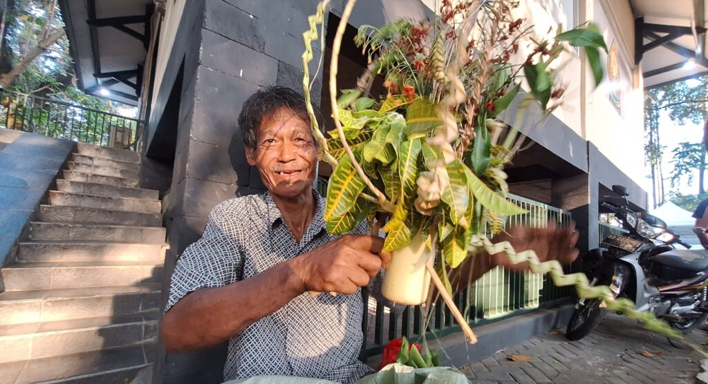 Screenshot_20260717_214236_Gallery - Mabur.co Smiling man presenting a vase of tropical leaves and flowers outdoors by a dark wall and stairs.