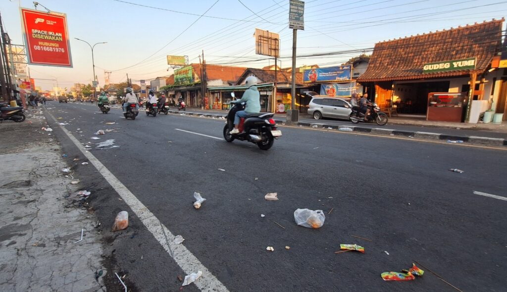 Street scene with several motorcycles riding on a litter-strewn city road in front of small shops and eateries at sunset/sunrise light.