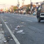 Urban street littered with trash along the curb while motorcycles and pedestrians line the sidewalks; a pickup truck drives by.