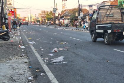 Urban street littered with trash along the curb while motorcycles and pedestrians line the sidewalks; a pickup truck drives by.