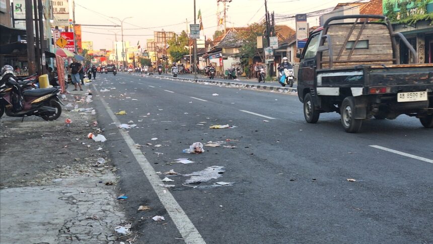 Urban street littered with trash along the curb while motorcycles and pedestrians line the sidewalks; a pickup truck drives by.