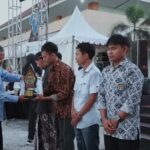 An older man in a light blue shirt hands a trophy to a man wearing a batik shirt during an outdoor award ceremony, with other recipients and onlookers in the background under a white tent and palm trees.