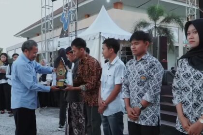 An older man in a light blue shirt hands a trophy to a man wearing a batik shirt during an outdoor award ceremony, with other recipients and onlookers in the background under a white tent and palm trees.