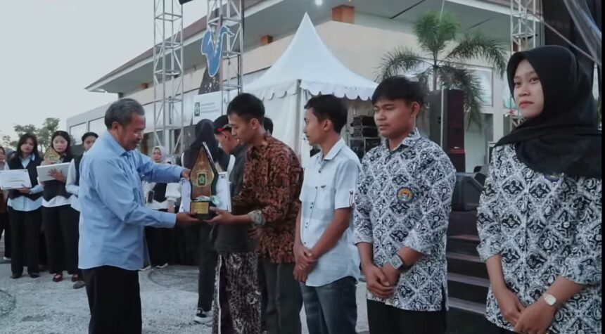 An older man in a light blue shirt hands a trophy to a man wearing a batik shirt during an outdoor award ceremony, with other recipients and onlookers in the background under a white tent and palm trees.