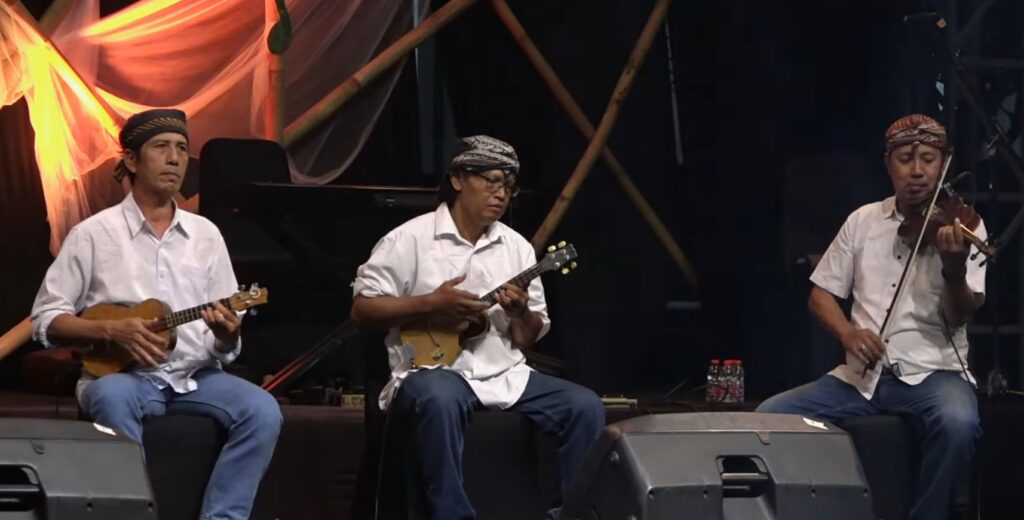 Screenshot_20260722_081258 - Mabur.co Three male musicians in white shirts and patterned headbands play stringed instruments on a stage.