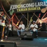 Male singer in a colorful patterned jacket sings into a microphone on stage as a five-member keroncong band plays behind him, with a banner reading 'KERONCONGLARAS TAMAN BUDAYA'.