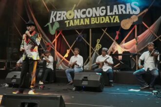 Male singer in a colorful patterned jacket sings into a microphone on stage as a five-member keroncong band plays behind him, with a banner reading 'KERONCONGLARAS TAMAN BUDAYA'.