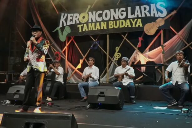 Male singer in a colorful patterned jacket sings into a microphone on stage as a five-member keroncong band plays behind him, with a banner reading 'KERONCONGLARAS TAMAN BUDAYA'.