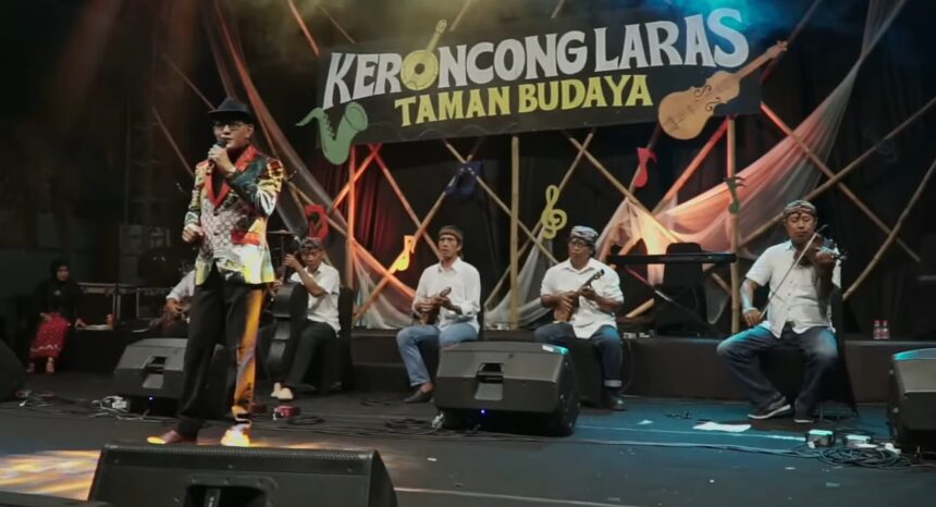 Male singer in a colorful patterned jacket sings into a microphone on stage as a five-member keroncong band plays behind him, with a banner reading 'KERONCONGLARAS TAMAN BUDAYA'.