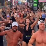 Large group of people wearing wireless headphones dancing and cheering in a busy street with colorful signage in the background (silent disco-style event).
