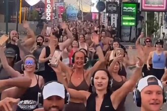 Large group of people wearing wireless headphones dancing and cheering in a busy street with colorful signage in the background (silent disco-style event).