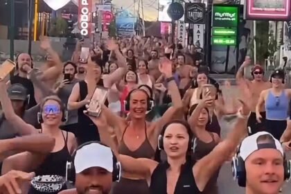 Large group of people wearing wireless headphones dancing and cheering in a busy street with colorful signage in the background (silent disco-style event).