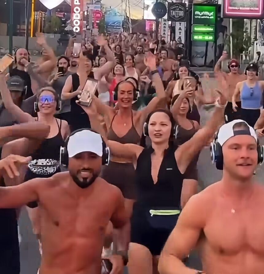 Large group of people wearing wireless headphones dancing and cheering in a busy street with colorful signage in the background (silent disco-style event).