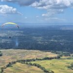 Paraglider with a yellow canopy gliding over patchwork farmland and distant hills under a blue sky.