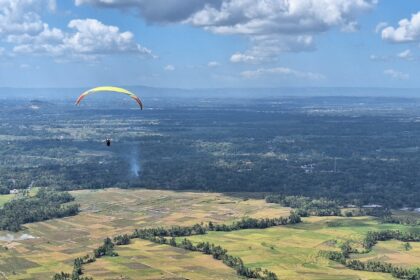 Paraglider with a yellow canopy gliding over patchwork farmland and distant hills under a blue sky.