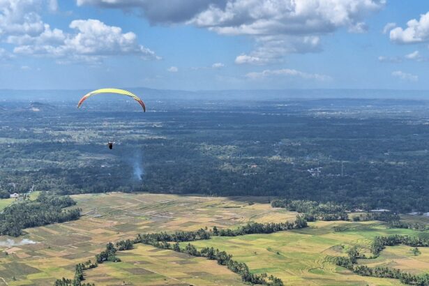Paraglider with a yellow canopy gliding over patchwork farmland and distant hills under a blue sky.