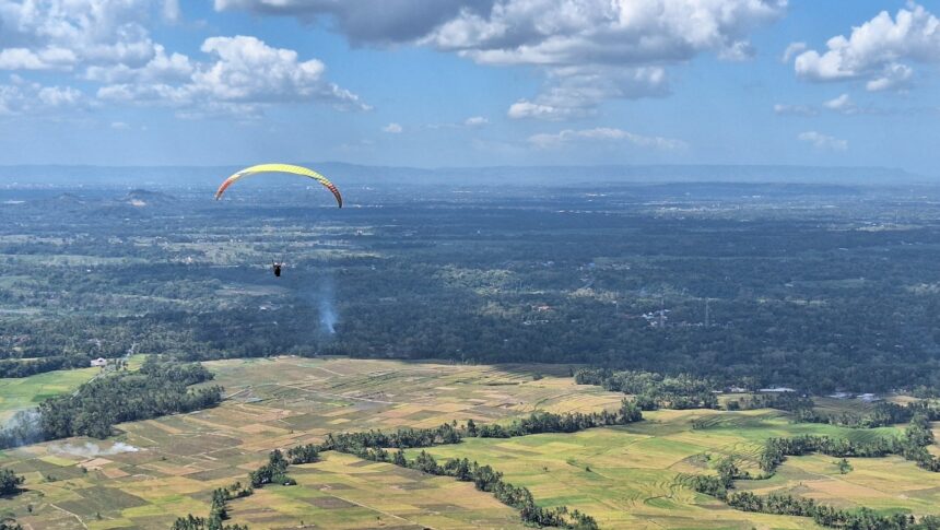 Paraglider with a yellow canopy gliding over patchwork farmland and distant hills under a blue sky.