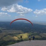 Red paraglider arcs across a blue sky over patchwork fields, with spectators on a hilltop viewing platform below.