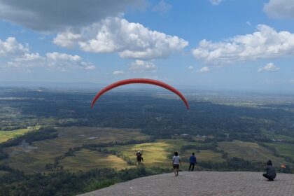 Red paraglider arcs across a blue sky over patchwork fields, with spectators on a hilltop viewing platform below.