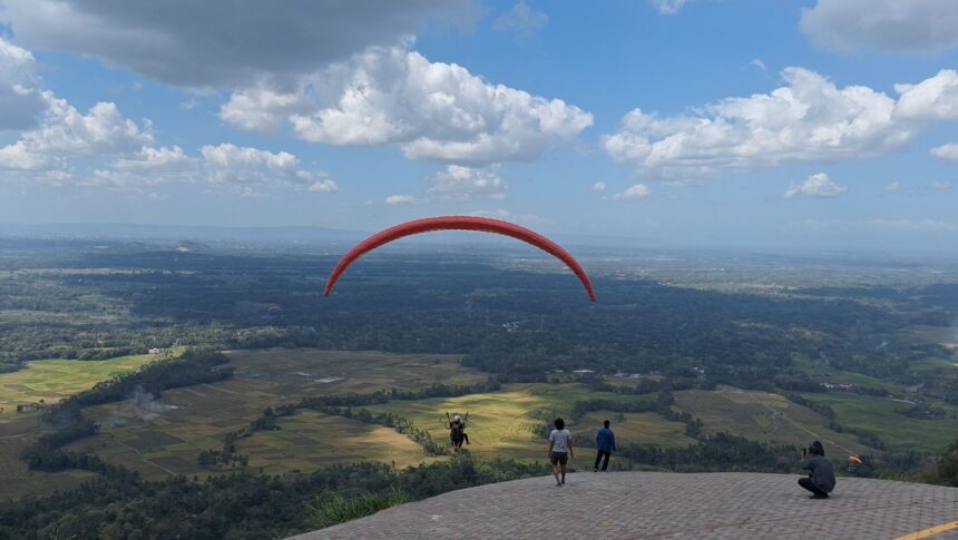 Red paraglider arcs across a blue sky over patchwork fields, with spectators on a hilltop viewing platform below.