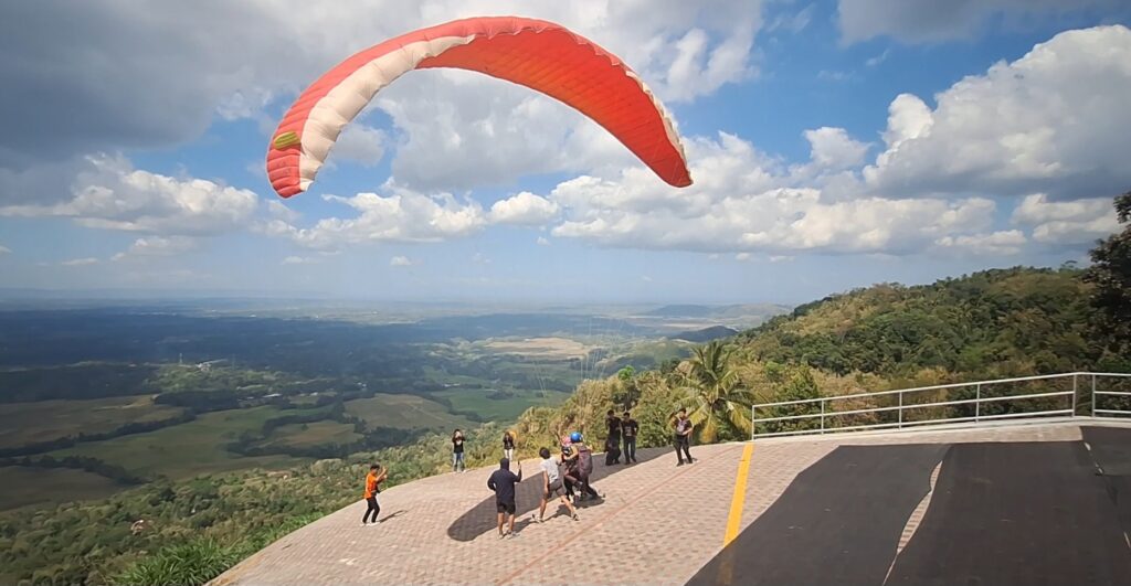 Paraglider in a red-and-white canopy lifts off from a hilltop launch platform as people watch nearby.