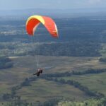 Paraglider with a red and yellow wing soaring over patchwork fields and distant hills.