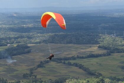 Paraglider with a red and yellow wing soaring over patchwork fields and distant hills.