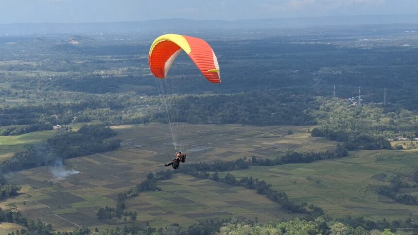 Paraglider with a red and yellow wing soaring over patchwork fields and distant hills.
