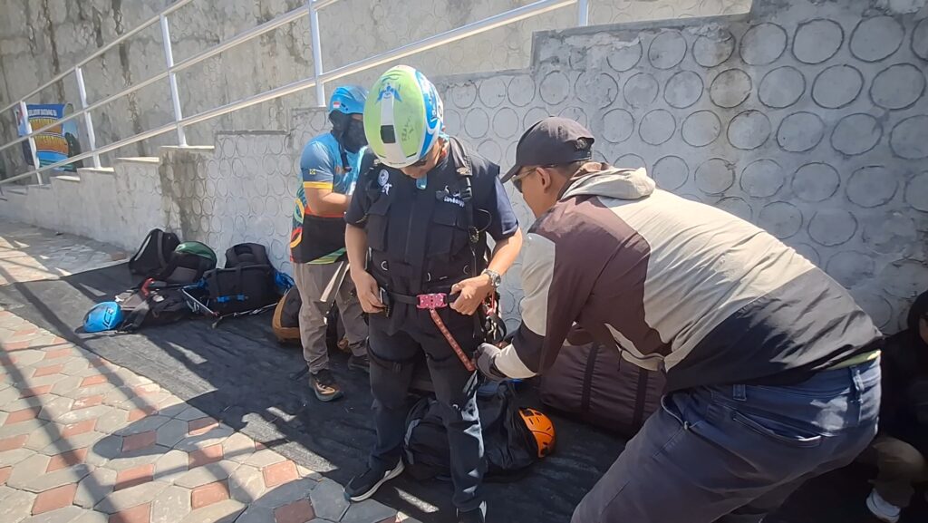 Climbers gather gear and secure a harness near a stone wall and stairs visible in the background.